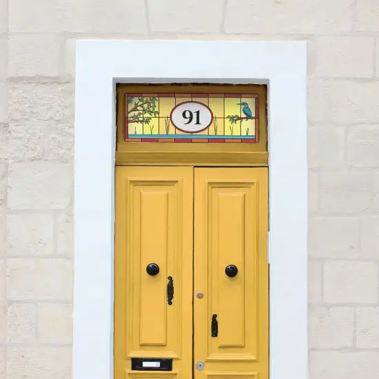 A modern stained glass window film design with a small bird and a central house number with red borders, installed above a traditional yellow door.